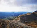 Un randonneur solitaire entame sa grande traversée des Alpes, dominant une vallée verdoyante sous un ciel nuageux.