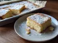 En bit fluffig, nybakad mjuk kaka med pudersocker på en grå tallrik. Resten av kakan syns i en ugnsform i bakgrunden.