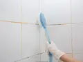Mano con guanto bianco usa spazzola blu per pulire le fughe delle piastrelle bianche, mostrando come togliere la muffa.