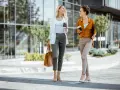 Dos mujeres con looks para ir a trabajar, tomando café y charlando, caminan frente a un edificio moderno.