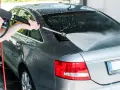 A person demonstrates how to power wash a car, spraying water onto the rear of a grey sedan.