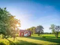Czerwony domek na wsi, otoczony zieloną trawą i drzewami. Słońce przebija się przez liście, tworząc długie cienie. To idealne miejsce, gdzie można odpocząć, z dala od miejskiego zgiełku. Oznaczenie użytku r.