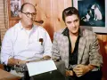Tom Parker und Elvis Presley sitzen nebeneinander. Parker, mit einer Pfeife im Mund, tippt auf einer Schreibmaschine.