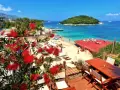 Albania camping nad morzem: czerwone kwiaty na tarasie z widokiem na piaszczystą plażę, turkusowe morze i zieloną wyspę.
