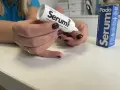 Dłonie z ciemnym lakierem aplikują podoserum na paznokieć. Opakowanie Podo Serum widoczne obok.
