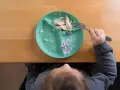 Dziecko je kurczaka z talerza babyono z przyssawką. Resztki jedzenia na talerzu i widoczna rączka z widelcem.