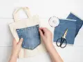 Mains épinglant une poche extérieure en jean sur un sac en toile. Ciseaux et chutes de tissu à proximité.