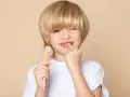 Chłopiec z blond włosami trzyma różową szczoteczkę, a palcem wskazuje na swoje zęby, sugerując krwawiące dziąsła u dziecka.