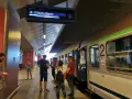 Ludzie na peronie czekają na pociąg, na tablicy informacyjnej widnieje godzina 16:12 i kierunek Gdynia Główna.