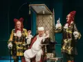 Chory z urojenia siedzi na tronie, otoczony przez dwóch dziwacznie ubranych mężczyzn w perukach.