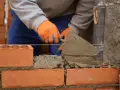Budowa murowanego prostego grilla z cegły. Ręce w pomarańczowych rękawiczkach nakładają zaprawę kielnią.