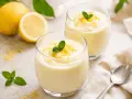 Due bicchieri di soffice mousse al limone, guarniti con scorza di limone e menta fresca. Sullo sfondo, limoni interi e tagliati.