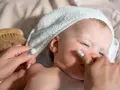 Niebieskooki noworodek w jasnym ręczniku na głowie, przypominającym czapkę. Delikatne dotknięcie palcem policzka.
