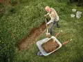 Mann gräbt einen Graben, um den Hang abzufangen. Erde wird in eine Schubkarre geschaufelt.