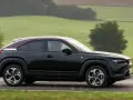 Czarna Mazda MX-30 z bordowym dachem sunie po drodze wśród zielonych wzgórz.