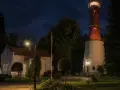 Nocą rozświetlona latarnia morska Rozewie strzeże wybrzeża. Obok niej stoją budynki i ceglany komin.