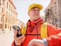 Kurier Glovo w żółtej czapce i pomarańczowej kurtce sprawdza telefon, gotowy do odbioru płatności Glovo cash.