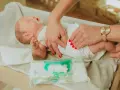 Delikatne dłonie z czerwonymi paznokciami pokazują, jak przewijać noworodka, używając chusteczek nawilżanych.