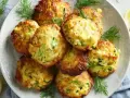 Goldbraune Zucchinipuffer, perfekt für Kinder, auf einem Teller mit Dill und Zitronenspalten garniert.