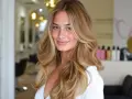 Uśmiechnięta blondynka z fryzurą butterfly cut, z falowanymi włosami w odcieniach blondu i brązu, prezentuje efektowną metamorfozę w salonie fryzjerskim.