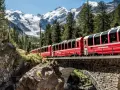 A red train travels across a stone bridge, winding its way from Chur to Tirano through a stunning alpine landscape with snow-capped mountains.