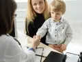 Mama z synkiem u lekarza. Chłopiec ściska dłoń lekarce, a mama uśmiecha się. W tle widać tablet i białą buteleczkę, jakby z lekiem. To ważny moment, być może wpis do książeczki zdrowia.