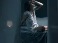 Frau sitzt im Bett, die Stirn gerunzelt, die Hand an der Stirn. Sie fragt sich, wie viel schwitzt man in der Nacht.