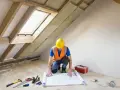 Budowlaniec w kasku i kamizelce analizuje projekt na podłodze w surowej suterenie. Narzędzia i skrzynka z wyposażeniem leżą obok.