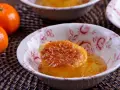 Un esponjoso baba al ron bañado en almíbar, adornado con ralladura de naranja, en un cuenco floral. Cerca, mandarinas frescas.