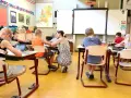 Dzieci siedzą w klasie, ucząc się. Nauczycielka pochyla się nad jednym z uczniów. Ile jest klas w Anglii? Tego nie wiemy, ale ta lekcja wygląda na interesującą.