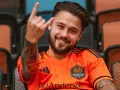 Uśmiechnięty Adam Kowalczyk w koszulce Houston Dynamo FC pozdrawia gestem rock and rolla, siedząc na trybunach.
