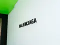 Il logo Balenciaga su una parete bianca, sotto un soffitto verde brillante. Chi è Balenciaga? Un nome che evoca alta moda e design audace.