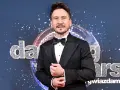 Tomasz Barański w czarnym smokingu pozuje na tle błyszczącej kuli dyskotekowej i napisu "Dancing with the Stars".