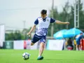 Piłkarz w białej koszulce z niebieskimi rękawami i spodenkami kopie piłkę na zielonym boisku.