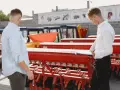Dwóch mężczyzn ogląda czerwoną maszynę rolniczą, prawdopodobnie siewnik lub bronę, na tle innych maszyn i budynków.