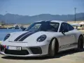 Szary Porsche 911 Carrera z czarnymi pasami na masce i dachu, zaparkowany nad wodą z górami w tle.