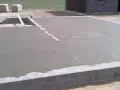 Świeżo wylany beton na chudziaka tworzy fundament pod budowę. W tle widać materiały budowlane i fragment budynku.