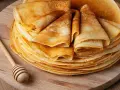 Pile de crêpes dorées, prêtes à être garnies. Une cuillère à miel en bois repose à côté, suggérant une délicieuse recette de crêpes.