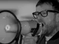 Un homme avec des lunettes et un mégaphone, sa voix résonne. Le groupe est en plein concert.