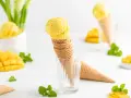 Zwei Kugeln cremiges Mango Eis in Waffelhörnchen, umgeben von frischen Mangostücken und Minze. Ein süßer Genuss!
