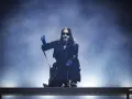 Jared Leto w niebieskich rękawiczkach, w okularach i długim płaszczu, klęczy na scenie. Jego zespół pewnie czeka na jego znak.