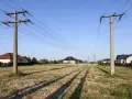 Linia średniego napięcia na działce, z której rozciągają się cienie na trawiastym terenie. W tle widać domy mieszkalne.