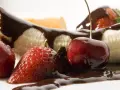 Soczyste truskawki i wiśnie oblane czekoladą, obok kawałków banana. Pyszne owoce w czekoladzie kuszą swoim wyglądem.