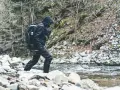 Wędkarz w czarnym ubraniu przechodzi przez kamienisty brzeg potoku, trzymając wędkę. W tle las i zbocze. Zalew Nakło Chechło.