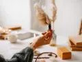 Mano sosteniendo un palo santo humeante, rodeada de velas, libros y cristales, creando un ambiente de sanación y paz.