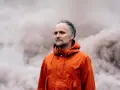Artur Rojek w pomarańczowej kurtce, otoczony mgłą. Jakby czekał na catchup, ale wokół pusty krajobraz. Magiera dymu.