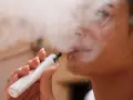 Una mujer exhala vapor de un vaper blanco. El vapor parece rodear su rostro, sugiriendo que el vaper encharca los pulmones.