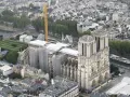 Widok z lotu ptaka na katedrę Notre Dame po pożarze, otoczoną rusztowaniami i placem budowy. Żuraw budowlany góruje nad okolicą.
