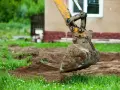 Błotnista łyżka koparki wykonuje niwelację terenu wokół domu.