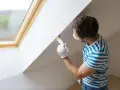 Mężczyzna w masce i rękawiczkach maluje pędzlem białą ścianę pod skosem. To malowanie ścian krok po kroku.